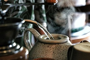 Graue Teekanne wird mit Wasser aufgegossen, vorne Links ist angeschnitten eine Kaffeemühle zu sehen.