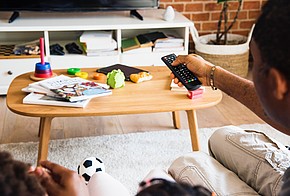 Man mit ausgestrecktem Arm und Fernbedienung in der Hand vor Couchtisch mit Broschüren darauf.